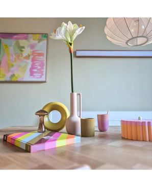 Ein moderner Holztisch zeigt farbenfrohe Objekte, darunter eine rosa Vase mit der Kunstblume Amaryllis Real Touch weiß 66 cm, eine gelbe Ringskulptur, gestreifte Bücher und eine rosa Tasse. Abstrakte Kunst akzentuiert die Hintergrundwand.