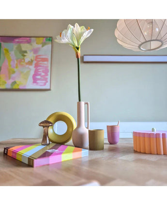 Ein moderner Holztisch zeigt farbenfrohe Objekte, darunter eine rosa Vase mit der Kunstblume Amaryllis Real Touch weiß 66 cm, eine gelbe Ringskulptur, gestreifte Bücher und eine rosa Tasse. Abstrakte Kunst akzentuiert die Hintergrundwand.
