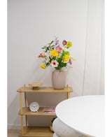 Auf einem hellen Holzregal stehen eine beige Vase mit künstlichen Ranunkeln in Real Touch Yellow (60 cm), eine weiße Uhr und ein Holztablett. Im Vordergrund erscheint ein Teil eines runden weißen Tisches vor einer schlichten weißen Wand.