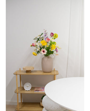 Auf einem hellen Holzregal stehen eine beige Vase mit künstlichen Ranunkeln in Real Touch Yellow (60 cm), eine weiße Uhr und ein Holztablett. Im Vordergrund erscheint ein Teil eines runden weißen Tisches vor einer schlichten weißen Wand.