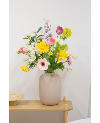 Auf einem hellen Holztisch vor einer schlichten weißen Wand steht eine beige Vase mit einer 60 cm großen Kunstblume „Artificial Ranunculus Real Touch Yellow“.