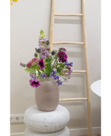 Eine 62 cm große, künstliche Glockenblume in Lila steht in einer Milchglasvase auf einem strukturierten weißen Sockel. Dahinter lehnt eine helle Holzleiter an einer beigen Wand, in Bodennähe sind Steckdosen sichtbar.