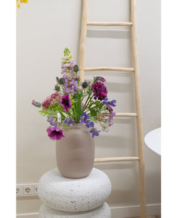 Eine 62 cm große, künstliche Glockenblume in Lila steht in einer Milchglasvase auf einem strukturierten weißen Sockel. Dahinter lehnt eine helle Holzleiter an einer beigen Wand, in Bodennähe sind Steckdosen sichtbar.