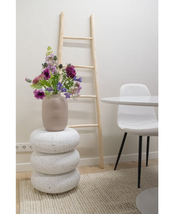 Eine künstliche Blumen-Glockenblume 62 cm lila steht in einer beigen Vase auf gestapelten weißen Terrazzohockern. Dahinter befindet sich eine Leiter aus hellem Holz, daneben ein weißer Stuhl und ein runder Tisch, alles auf einem strukturierten beigen Teppich in einem minimalistischen Raum platziert.