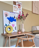 Ein Holzschreibtisch mit passendem Stuhl zeigt einen blauen Vogelaufdruck, eine künstliche Blume Anemone verzweigt 56 cm rosa, ein rundes gelbes Ornament und eine kleine Tasse. Oben hängt neben einer beigen Couch eine Pinnwand mit Fotos und Notizen.