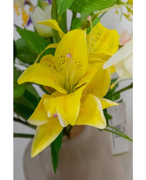 Eine Nahaufnahme der Kunstblume Asiatische Lilie 66 cm gelb, mit leuchtend gelben Blütenblättern mit dunklen Sprenkeln, umgeben von grünen Blättern und anderen Blumen in einer hellen Vase.