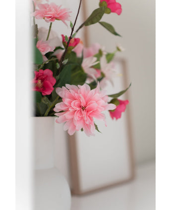 Eine Nahaufnahme der Kunstblumen-Dahlie 68 cm rosa in einer weißen Vase, mit einem schwachen Fotorahmen im Hintergrund auf einer weißen Oberfläche.