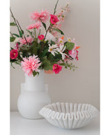 Eine Kunstblumen-Dahlie 68 cm rosa in einer weißen Vase steht neben einer weißen Keramikschale mit Rüschen auf einer weißen Fläche vor einem schlichten hellen Hintergrund.