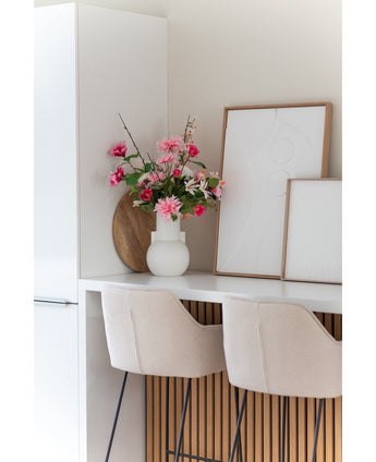 Eine moderne weiße Küche mit zwei beigen Barhockern, einer 68 cm großen künstlichen Dahlie in Rosa in einer weißen Vase auf der Theke und zwei gerahmten abstrakten Kunstwerken an der Wand.