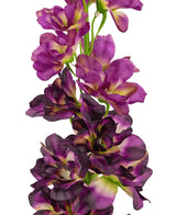 Eine Nahaufnahme der künstlichen Blume Delphinium Purple 79 cm, ein hoher künstlicher Stiel mit mehreren violetten und dunkelvioletten Blüten mit einem grünen und gelben Herzen, vor einem weißen Hintergrund.