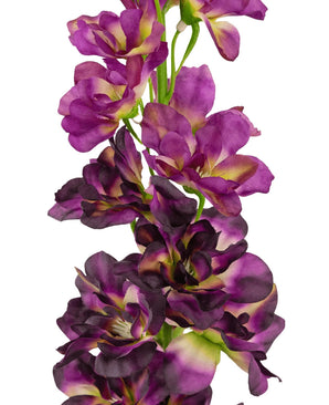 Eine Nahaufnahme der künstlichen Blume Delphinium Purple 79 cm, ein hoher künstlicher Stiel mit mehreren violetten und dunkelvioletten Blüten mit einem grünen und gelben Herzen, vor einem weißen Hintergrund.