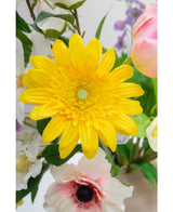 Die Kunstblume Gerbera 53 cm gelb, eine leuchtend gelbe Kunstblume mit geschichteten Blütenblättern, steht zwischen anderen farbenfrohen Kunstblumen und grünen Blättern, mit weißen und rosa Blüten im Hintergrund.