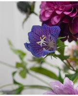 Eine Nahaufnahme der Kunstblume Harebell 62 cm lila, einer zarten lila Kunstblume mit tiefen Adern und gelben Staubgefäßen, zwischen grünen Blättern und zarten zartrosa und violetten Blüten im Hintergrund.