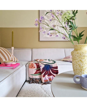 Ein gemütliches Wohnzimmer mit einem hellbeigen Sofa, einem geblümten Plaid, einer rosa Kerze, einem gestreiften Kissen und einer künstlichen Spitzenblume 76 cm weiß in einer gelben Vase auf dem runden Tisch. Abstrakte Kunst schmückt die Wand dahinter.