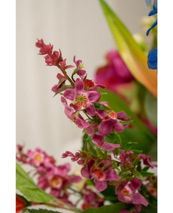 Nahaufnahme der künstlichen Blumenkatzenminze 75 cm rosa: eine Gruppe leuchtend rosafarbener Kunstblumen mit einem grünen Herzen und üppigen Blättern, scharf dargestellt vor einem sanft verschwommenen Hintergrund mit Andeutungen anderer Blumen.