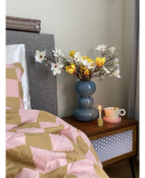 Ein Nachttisch mit einer blauen Vase mit der Kunstblume Magnolie 50 cm gelb/orange, einer rosa Teetasse auf einer gänseblümchenförmigen Untertasse und einer brennenden Kerze steht neben einem Bett, das mit einer rosa-braunen Decke mit geometrischen Mustern bedeckt ist.