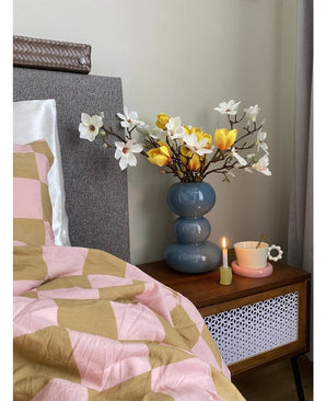 Ein Nachttisch mit einer blauen Vase mit der Kunstblume Magnolie 50 cm gelb/orange, einer rosa Teetasse auf einer gänseblümchenförmigen Untertasse und einer brennenden Kerze steht neben einem Bett, das mit einer rosa-braunen Decke mit geometrischen Mustern bedeckt ist.