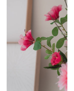 Eine Nahaufnahme der künstlichen Blume Rhododendron Azalee 73 cm fuchsia, mit leuchtend rosa künstlichen Blumen mit grünen Blättern, vor einem weichen, neutralen Hintergrund und einem Teil eines Holzrahmens sichtbar.