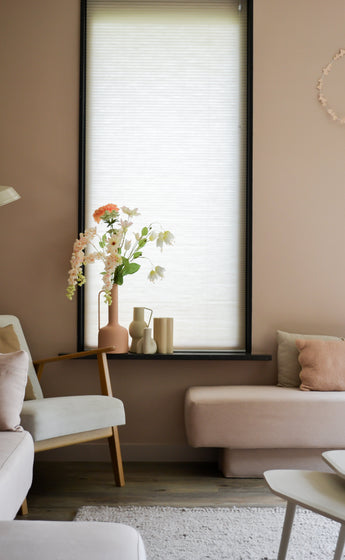 Ein gemütliches Wohnzimmer mit hellrosa Wänden, einem großen Fenster mit einem weißen Rollo, cremefarbenen und rosafarbenen Sofas und einem Holzstuhl mit der künstlichen Blumen-Perlmutterfalter 94 cm weiß neben dekorativen Objekten auf der Fensterbank.