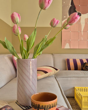Eine 63 cm große künstliche französische Tulpe in Rosa steht in einer Vase auf dem Tisch neben einer orangefarbenen Tasse in einem gemütlichen Wohnzimmer mit einem cremefarbenen Sofa, gemusterten Kissen, einer gelben Kiste und abstrakter Wandkunst im Hintergrund.