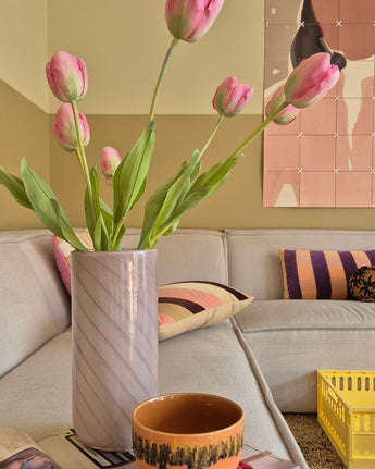Eine 63 cm große künstliche französische Tulpe in Rosa steht in einer Vase auf dem Tisch neben einer orangefarbenen Tasse in einem gemütlichen Wohnzimmer mit einem cremefarbenen Sofa, gemusterten Kissen, einer gelben Kiste und abstrakter Wandkunst im Hintergrund.