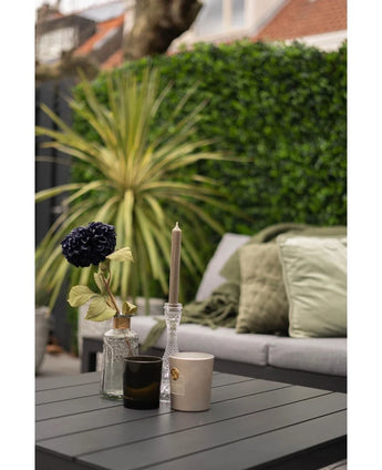Ein moderner Outdoor-Tisch mit schwarzer Oberfläche zeigt eine Vase mit einer dunklen Blume, einen Kerzenhalter aus Glas und zwei Tassen. Dahinter steht ein Sofa mit grünen Kissen gegenüber einer üppig grünen Wand aus dem Kunsthaag-Mattenmix 50x50 cm.