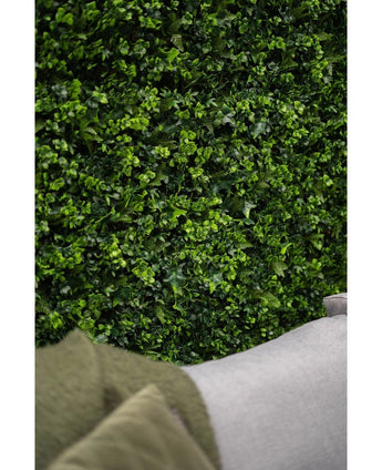 Nahaufnahme einer üppig grünen Wand aus künstlichem Heckenmattenmix im Format 50 x 50 cm mit leuchtenden Blättern, wobei im unteren Vordergrund die Ecke eines hellgrauen Sofas und ein Teil eines grünen Mantels zu sehen sind.