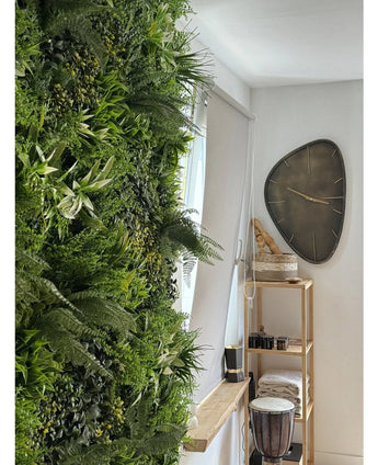 Eine üppige künstliche Hecke Freshness 100x100 cm UV-Vertikalgarten bedeckt eine Wand eines hellen Raumes. Daneben hängt eine moderne dreieckige Wanduhr über einem Holzregal mit Handtüchern, einer Djembe-Trommel, Körben und kleinen Dekostücken.