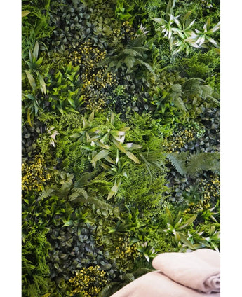 Eine üppige, strukturierte Kulisse wird durch die vertikale UV-Gartenwand Kunsthaag Freshness 100 x 100 cm geschaffen, die dicht mit grünem Blattwerk bedeckt ist. Unten rechts sind zwei gefaltete beige Handtücher zu sehen.