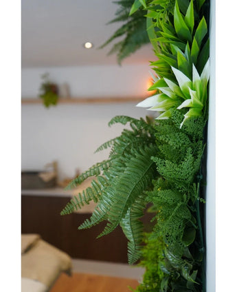 Eine Nahaufnahme üppig grüner künstlicher UV-Hedge Freshness 100x100 cm ziert eine vertikale Innenwand, mit einem verschwommenen Hintergrund, der neutrale Möbel und eine kleine Hängepflanze zeigt.