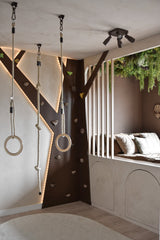 Ein modernes Kinderspielzimmer verfügt über eine Kletterwand, Turnringe, Seil und eine gemütliche Leseecke mit Kissen und vertikalen Lattenrosten. Die künstliche Hecke Lush Deco Deluxe 100x100 cm UV bringt Grün an die Decke und ergänzt die neutralen Töne und die verspielte Beleuchtung des Raumes.