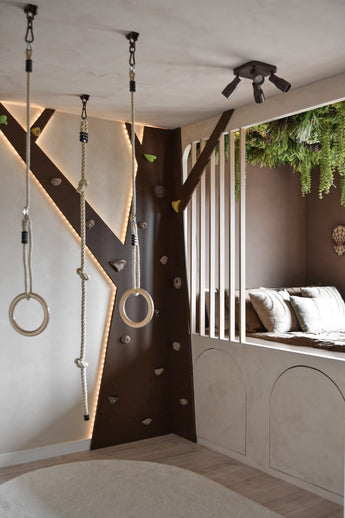 Ein modernes Kinderspielzimmer verfügt über eine Kletterwand, Turnringe, Seil und eine gemütliche Leseecke mit Kissen und vertikalen Lattenrosten. Die künstliche Hecke Lush Deco Deluxe 100x100 cm UV bringt Grün an die Decke und ergänzt die neutralen Töne und die verspielte Beleuchtung des Raumes.