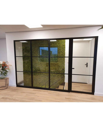 Ein modernes, luxuriöses Interieur mit einer schwarzen Glaswand offenbart eine grüne Wand aus künstlichem Haager Nordisches Moos 50x50 cm UV, eine Treppe dahinter, helle Holzböden und ein stilvolles Blumenarrangement auf der linken Seite.
