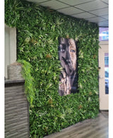 Eine 100 x 100 cm große feuerhemmende Kunsthaag Savanne-Platte mit üppigem Grün umgibt ein großes schwarz-weißes Gesichtsporträt in einem Innenraum mit gefliesten Deckenpaneelen und einer kleinen Theke auf der linken Seite.