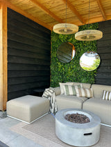 Moderner Sitzbereich im Freien mit beigem Sofa, gestreiften Kissen und rundem Feuertisch. Zwei runde Spiegel hängen an einer künstlichen Hecke Volta 100x100 cm UV-Pflanzenwand unter einer Holzdecke und Hängelampen aus Korbgeflecht.
