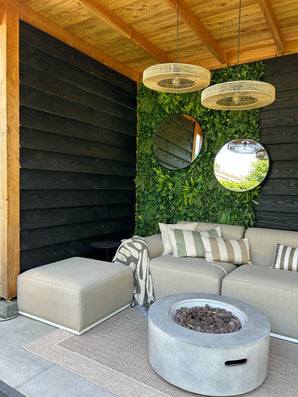 Moderner Sitzbereich im Freien mit beigem Sofa, gestreiften Kissen und rundem Feuertisch. Zwei runde Spiegel hängen an einer künstlichen Hecke Volta 100x100 cm UV-Pflanzenwand unter einer Holzdecke und Hängelampen aus Korbgeflecht.
