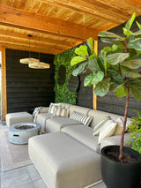 Ein gemütlicher Sitzbereich im Freien mit einem beigen Sofa, gestreiften Kissen, einer runden Feuerstelle, einer großen Topfpflanze und einer Holzpergola. Die Wand ist mit dem künstlichen Haag Volta 100x100 cm UV für zusätzlichen Stil mit Grün dekoriert.