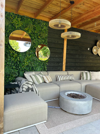 Eine gemütliche Terrasse mit beigem Sofa, gestreiften Kissen, Feuerstelle und gewebten Teppichen. UV-Grün von Kunsthaag Volta 100x100 cm hängt an der Wand, runde Spiegel und Korblampen unter einer Holzpergola.