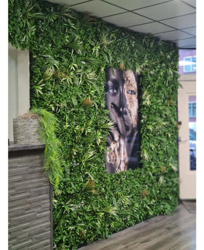 Eine mit künstlicher Heckenmatte Savanne 100x100 cm bedeckte Wand, mit UV-geschütztem Grün in einem Kunststoffpflanzgefäß Ø100, hat in der Mitte ein großes schwarz-weißes Porträt. Der moderne Innenraum verfügt über eine geflieste Decke und eine Holztheke auf der linken Seite.