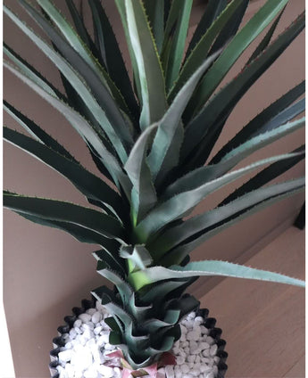 Eine Nahaufnahme der Kunstpflanze Agave Pandora UV 120 cm mit grünen, stacheligen Blättern in einem schwarzen Topf mit weißen Kieselsteinen, ausgestellt auf einer hellen Oberfläche vor einer neutralen Wand.