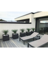 Auf einer modernen Terrasse stehen zwei graue Liegestühle und fünf quadratische Pflanzgefäße, jeweils mit einer künstlichen Pflanze Agave Pandora UV 85 cm, vor einer weiß-schwarz getäfelten Wand für einen eleganten, minimalistischen Look.