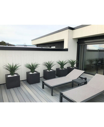 Auf einer modernen Terrasse stehen zwei graue Liegestühle und fünf quadratische Pflanzgefäße, jeweils mit einer künstlichen Pflanze Agave Pandora UV 85 cm, vor einer weiß-schwarz getäfelten Wand für einen eleganten, minimalistischen Look.