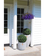 Auf der weißen Veranda eines Hauses befinden sich zwei UV-Topiary-Pflanzgefäße im römischen Stil (65 cm) mit künstlichem Buchsbaum auf Stiel und grünen Sträuchern, darüber ein Hängekorb mit violetten Blumen, weiße Säulen und eine Glastür auf der linken Seite.