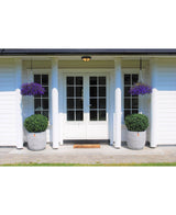 Weiße Doppeltüren mit Glasscheiben zieren ein weißes Vier-Säulen-Haus. Zwei künstliche Buchsbaumstiel UV D60 cm Topiaries und zwei hängende Körbe mit lila Blüten schmücken den Eingang, mit einer Fußmatte vor den Türen.