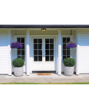 Weiße Doppeltüren mit Glasscheiben zieren ein weißes Vier-Säulen-Haus. Zwei künstliche Buchsbaumstiel UV D60 cm Topiaries und zwei hängende Körbe mit lila Blüten schmücken den Eingang, mit einer Fußmatte vor den Türen.