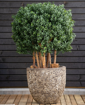 Die künstliche Buchsbaumkugel im römischen Stil UV 90 cm, eine runde grüne Blattpflanze mit dicken Stielen, steht in einem großen Steintopf auf Holz, vor einer dunklen horizontalen Wand mit Holzplatten.