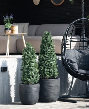 Zwei künstliche Buchsbaumpyramiden in UV-Form (90 cm) stehen auf einer Terrasse in der Nähe einer Außenbank und eines runden Hängesessels aus Korbgeflecht mit schwarzem Kissen, während im Hintergrund ein kleiner Tisch mit einer Pflanze steht.