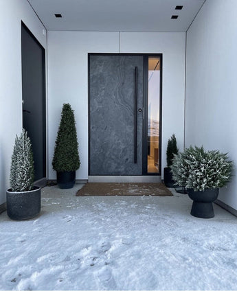 Eine moderne dunkle Haustür wird von weißen Wänden eingerahmt und flankiert von einer künstlichen Buchsbaumpyramide UV 90 cm, die für UV-Schutz sorgt. Leichter Schnee bedeckt den Boden, während diese stilvollen Gartenpflanzen einen aufgeräumten, minimalistischen Eingangsbereich schaffen.