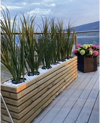 Auf einem Holzdeck steht ein Holzpflanzgefäß mit Kunstrasen Grün UV 90 cm und weißen Steinen neben einem schwarzen Pflanzgefäß mit bunten Hortensien, mit Glasbalustraden und Blick auf die Berge im Hintergrund.
