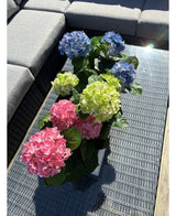 Die künstliche Hortensie Green UV 35 cm steht auf einem Korbtisch mit Glasplatte neben einem grauen Outdoor-Sofa mit realistischen grünen Hortensien, die im Sonnenlicht leuchten.
